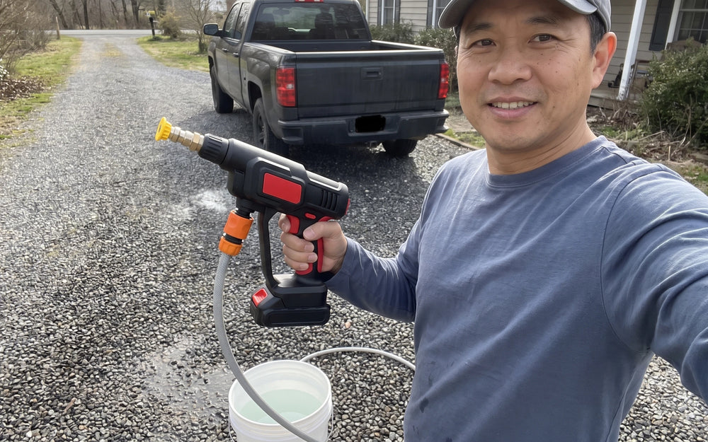 A smiling middle-aged Asian-American man in a slate-blue shirt and grey cap on a long gravel driveway holding a black-and-red cordless pressure washer in one hand, a white bucket of water at the feet of the man with a grey hose running into it and a dark pickup truck parked far up the drive behind the man