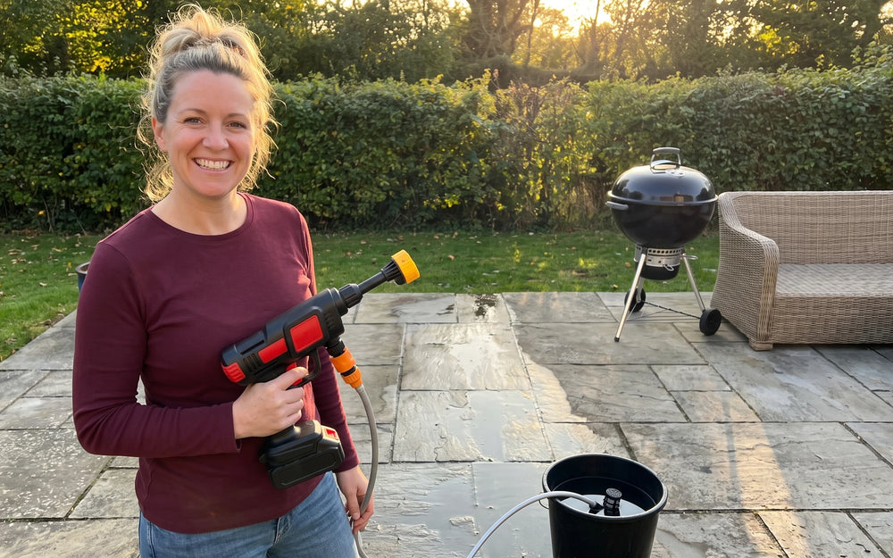 A stone patio in warm low afternoon sun, a smiling white woman in a burgundy shirt and jeans resting a black-and-red cordless pressure washer against one hip, one wide wet stripe across the grey flagstones in front of the woman, a black kettle barbecue and a wicker sofa at the right